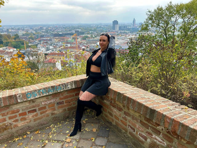 MayaCruise-hot with black leather jacket, black mini-skirt, fishnet stockings, and knee-high boots, sitting on brick wall, cityscape in background, overcast sky, fall leaves on ground.