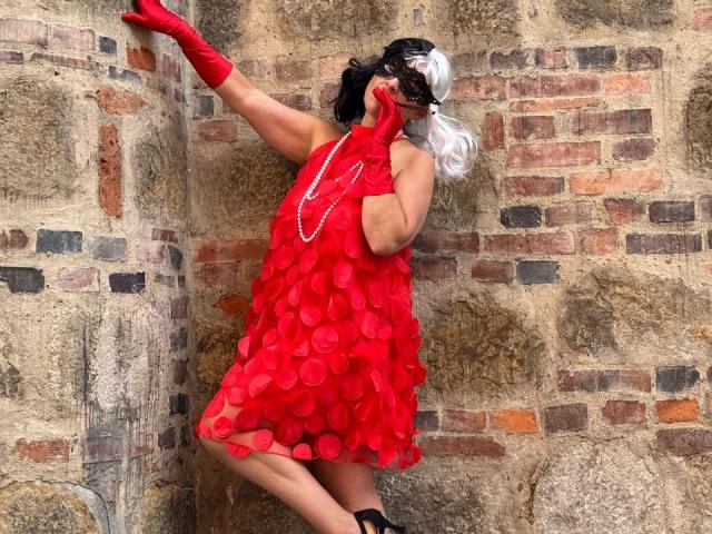 JaneTurner with black and white wig, red dress with circular patterns, red gloves, pearl necklace, and black heels, touching brick wall, holding mask to face, white-haired.
