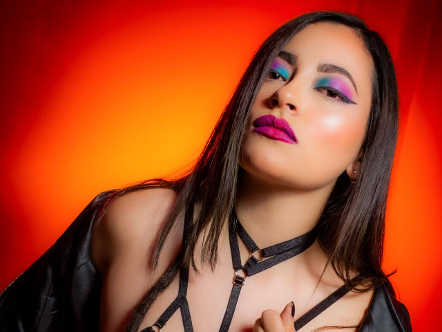 LunaTikka has pink lipstick, blue-green eyeshadow, and black strappy lingerie with metal rings, against a fiery red-orange background. She's looking up with a slight, confident smile.