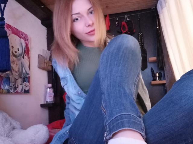 HarleyQuinn69 con cabello rubio, una blusa verde y jeans azules, con las piernas levantadas, mostrando ropa interior blanca, en una habitación de madera, mirando a la cámara con una ligera sonrisa..