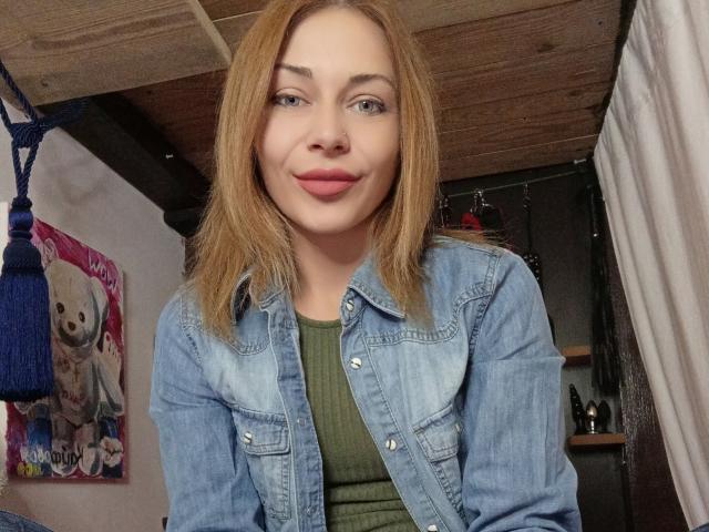 HarleyQuinn69 with light brown hair, green ribbed shirt, denim jacket, and pink lips, smiling slightly, nose ring, and blue eyes, in a wooden room with a blue tassel.