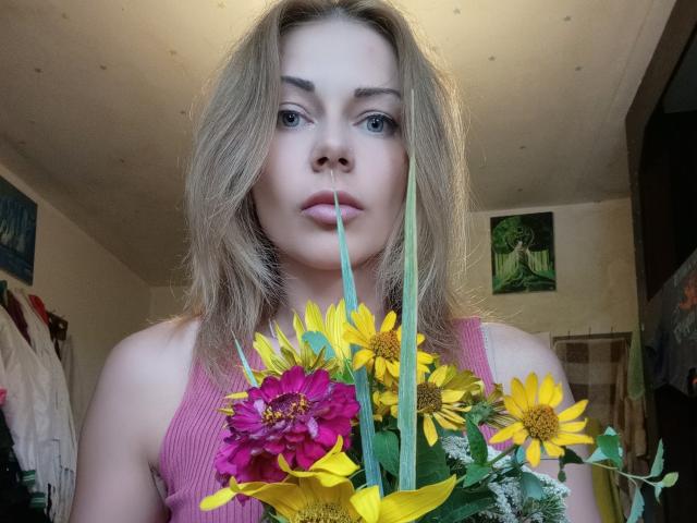 HarleyQuinn69 with blonde hair, pink tank top, holding a bouquet of yellow and pink flowers, green stem in mouth, serious expression, bedroom, soft lighting, blue eyes, fair skin.