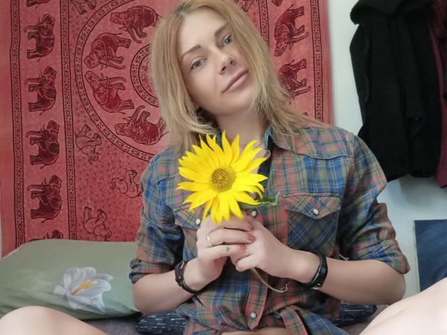 HarleyQuinn69 con i capelli biondi, che tiene un girasole giallo, indossa una camicia a quadri, è seduta su un letto, sorride, indossa un braccialetto nero e ha un anello alla mano destra..