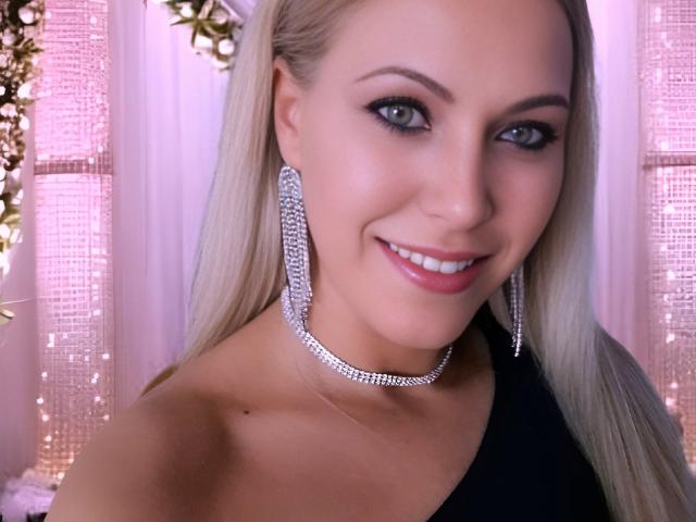 MyaHott tem cabelo loiro, olhos azuis e está sorrindo para a câmera. Ela está usando brincos de diamantes grandes e um colar combinando. Ela está vestida com uma roupa preta de um ombro só..