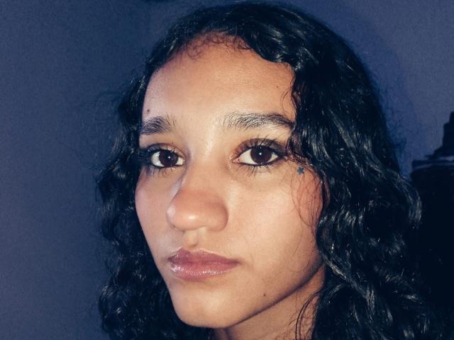 NikiSeexy heeft krullend zwart haar, bruine ogen en een schattig, lichtelijk fronsend gezicht met glanzende, roze lippen. Haar huid is glad en bruin. Ze ziet er serieus en aantrekkelijk uit..