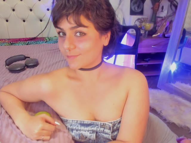 IsabelaCooper with short brown hair, nose ring, black choker, strapless denim top, holding apple, sitting on bed, smiling, visible tits, holding glasses in background.