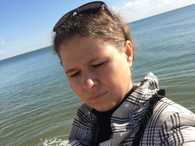 CurlyKsu with brown hair, sunglasses on head, plaid shirt, and black top, eyes closed, face showing discomfort, standing by a blue-green ocean under a clear sky.
