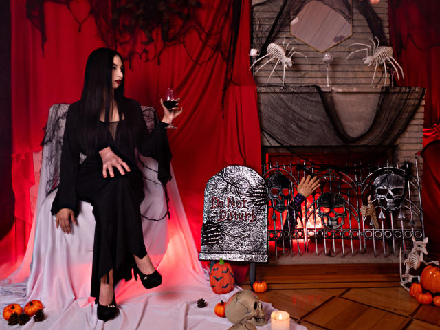 Camille Rios, com longos cabelos pretos, vestindo um vestido preto e saltos altos, sentada em uma cadeira, segurando um copo de vinho tinto, em um cenário de Halloween assustador, com abóbora, caveira e ossos..