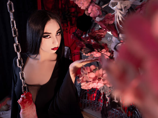 CamilleRios with dark hair, red lipstick, and black dress, holding chain with red hand, looking at pink flowers, in goth-style room, photo.