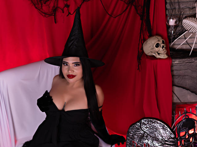 OliviGrace with black witch hat, red lipstick, black dress, showing her big boobs, sitting on white couch, black gloves, skull on red table, red and black decor.