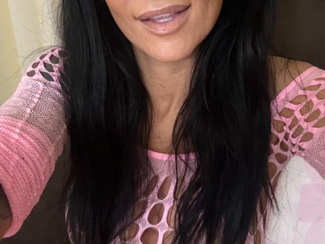 AndraD with long black hair, pink mesh top revealing her tits, and glossy pink lipstick, smiling slightly, showing her upper chest and mouth.