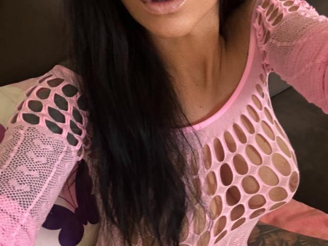 AndraD has black hair, pink fishnet top, and visible tits. Her lips are slightly parted, and she's wearing a pink lace garment. Her skin is light brown.