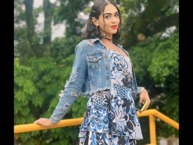 SexyHannaD with medium tits, wearing a blue denim jacket and patterned dress, standing by a yellow railing, holding a yellow object, green forest background.
