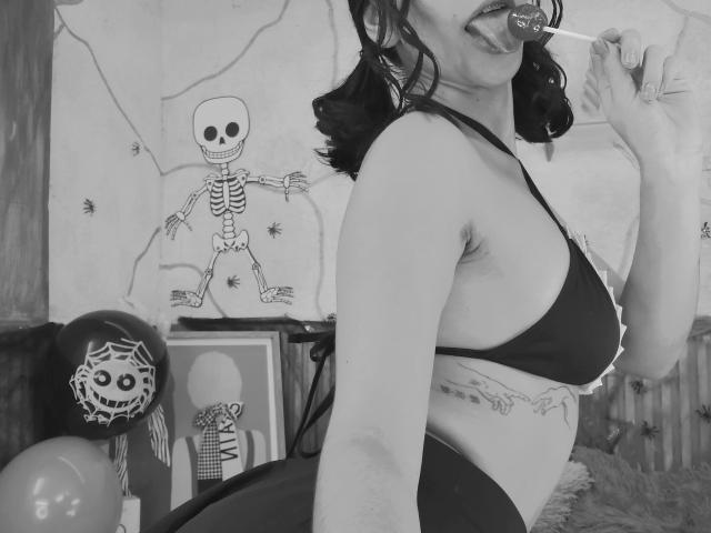 Black-and-white photo of LindaWatsong with dark wavy hair, sucking a lollipop, wearing a dark bra, showing her tits and a tattoo on her side. Skeleton drawing in background.