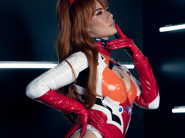 AlmaBella in a shiny red and white latex costume, with red gloves, pointing to her chin. She has long brown hair, a red headband, and a cleavage-cutout top.