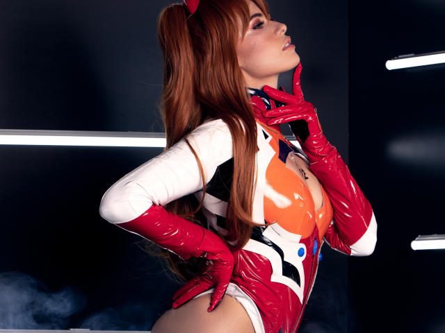 AlmaBella in a red and white latex bodysuit with red gloves, long red hair, and red headpiece, posing with fingers near her mouth, showing her tits.