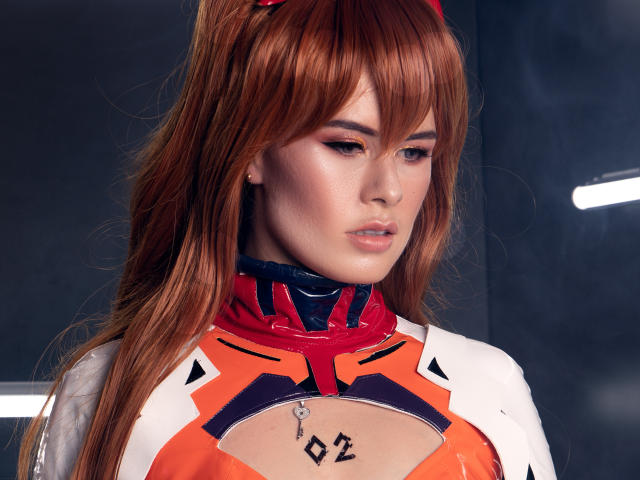 AlmaBella with red hair, wearing an orange and white shiny cosplay outfit, showing small tattoos on her chest, looking down with a serious expression. Boobs partially visible.