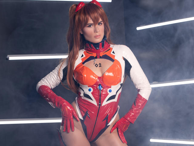 AlmaBella in a shiny red, white, and orange latex cosplay with red gloves, long brown hair in pigtails, and a red headpiece, standing with hands on hips.