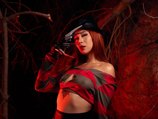 KatMore with red hair, black hat, and striped shirt, holding knife to her mouth, showing her bare tits and a tattoo on her belly, in dark red lighting.