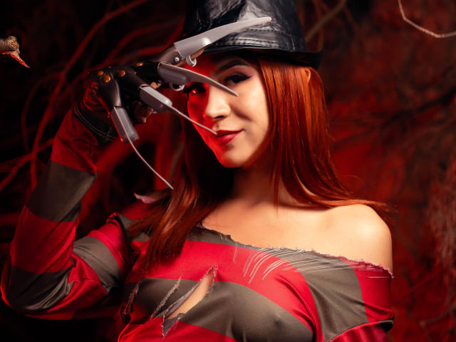 KatMore with red hair, wearing a black hat and striped red and gray shirt, holding sharp claws to her eye, smiling slightly, showing her bare shoulders.