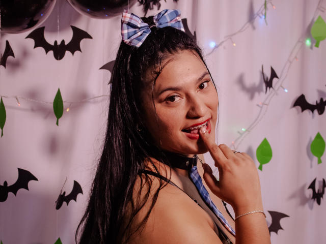 KorinaKova with dark hair in a ponytail, wearing a blue checkered bow, black choker, and sleeveless top, biting her finger, smiling, with bat decorations and string lights.
