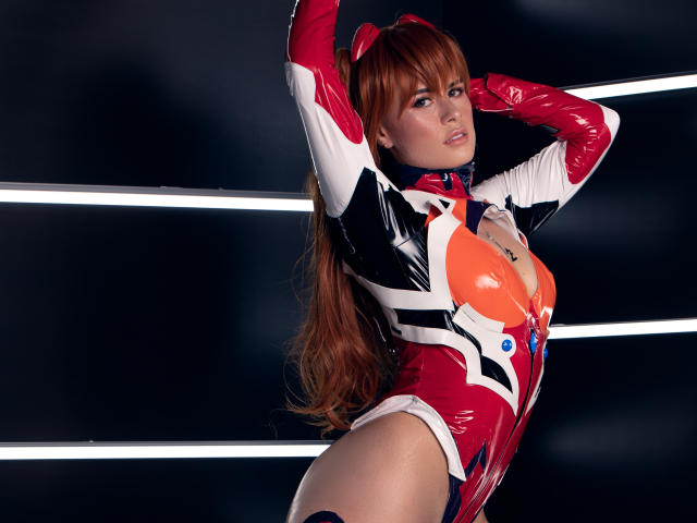 AlmaBella with long red hair, wearing a shiny red, white, and black latex bodysuit, arms raised, showing her orange boob cutout, and confident pose. Her pussy is not visible.