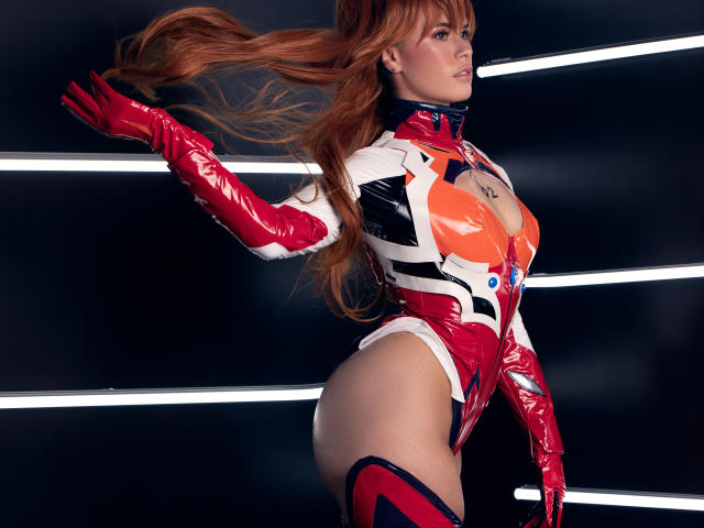 AlmaBella in a shiny red and white latex bodysuit with red gloves, showing big tits, a small ass, and long red hair, against a dark background with neon lines.