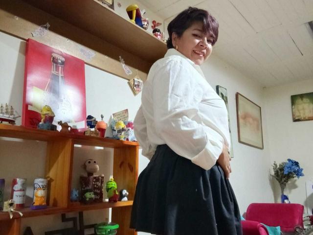 angellovelly con cabello castaño corto, sonriendo, vistiendo una camisa blanca, falda negra, de pie, techo blanco, estanterías de madera, juguetes pequeños, pintura roja, cuadros enmarcados, pared blanca, silla roja..
