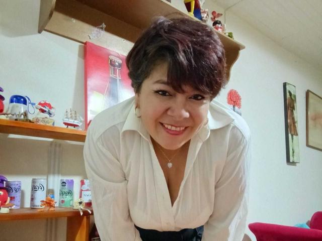 Angellovelly with short brown hair, smiling, wearing a white button-up shirt, black belt, and necklace, leaning forward, showing her small tits and cute face.