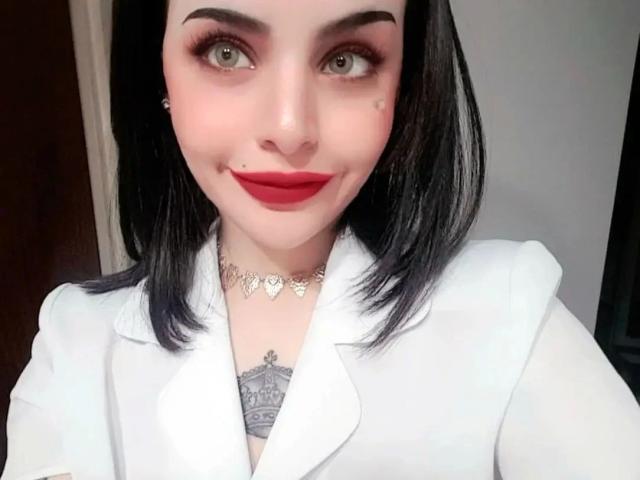 Celestia-hot with pale skin, black shoulder-length hair, red lipstick, white blazer, heart necklace, and tattoo under her chin, smiling with green eyes.