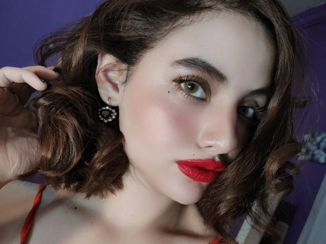 Celestia-hot with red lipstick, brown wavy hair, green eyes, and a small nose piercing, wearing red earrings, touching her hair, and a red strap visible.
