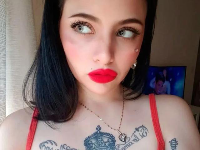 Celestia-hot with black hair, red lipstick, and tattoos on her chest, wearing a red top, has a nose piercing and a small facial stud, looking to the side.