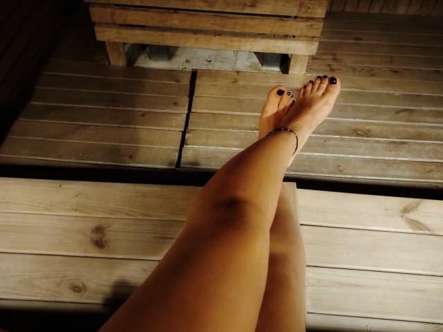 MistressMica's bare legs and feet with black-painted toenails crossed at the ankles, a wooden bracelet on her right ankle, on a wooden floor.