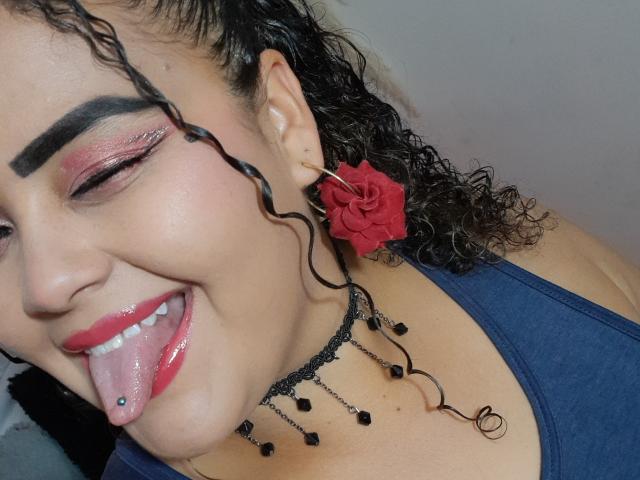 MelannieCarter has curly black hair, red lipstick, and a tongue piercing with a blue ball, wearing a red flower earring and black chain necklace, smiling with pink eyeshadow.