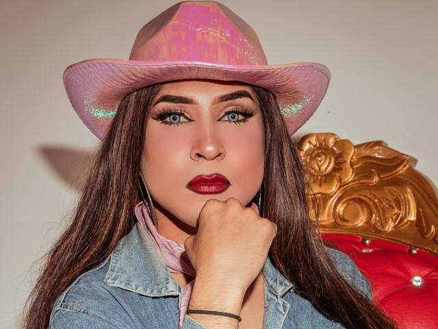 DarianaGio con cappello da cowboy rosa glitterato, occhi azzurri, lunghi capelli castani, rossetto rosso, giacca di jeans, sciarpa rosa, mano sul mento, sedia dorata decorata, cuscino rosso..