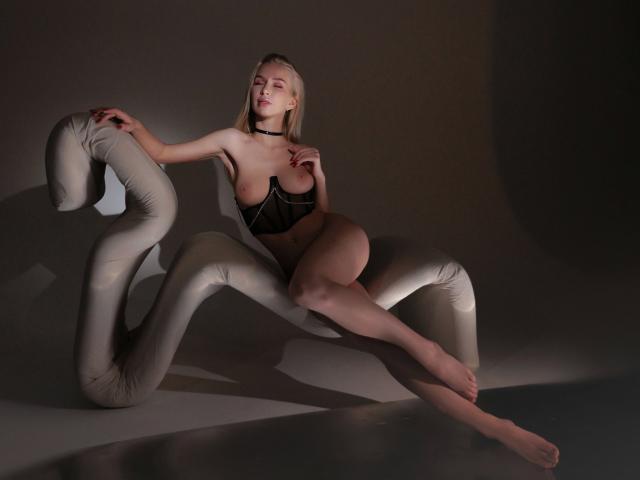 BlondiAngel-hot with bare boobs, wearing black lingerie, sits on a white, curly, inflatable chair, legs crossed, choker, dim lighting, shadows, and a sexy, confident expression.