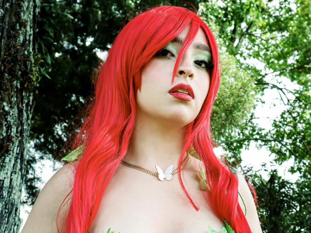 EvaMcAllister with bright red hair, green eye makeup, and a white butterfly necklace, looking up with red lips, her bare shoulders visible, standing in a green forest.