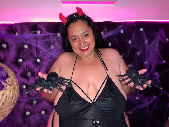 Latina, de complexión robusta, con cabello negro y cuernos de diablo rojos, vestida con un top negro de tirantes que deja ver sus grandes senos, sosteniendo guantes de araña negros, sonriendo, en un ambiente temático de Halloween..