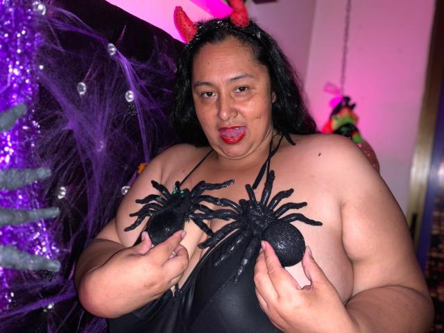 TistMature, a chubby woman with black hair and red devil horns, is holding black spider decorations over her large tits. She's sticking her tongue out slightly.