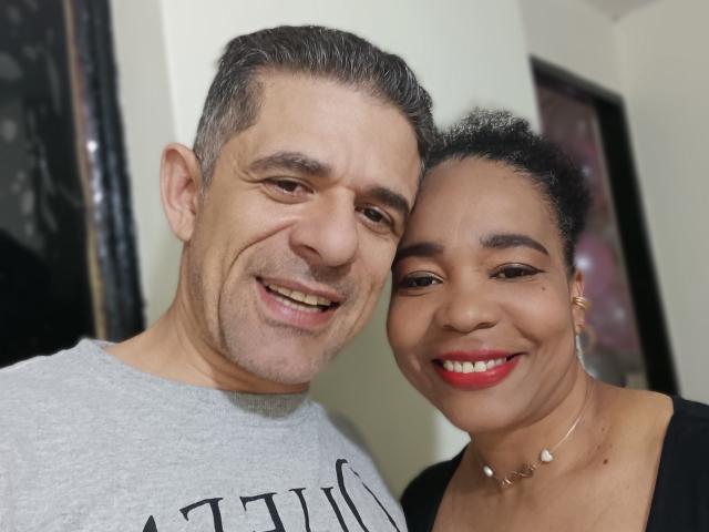 FernandoAndIsabella are smiling in a selfie. Fernando has gray hair and a beard, wearing a gray shirt. Isabella has curly black hair, red lipstick, and a black top.