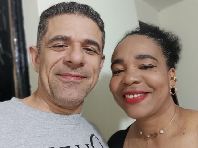 FernandoAndIsabella: smiling couple, Fernando with gray hair, slight beard, wearing gray shirt, Isabella with black hair in bun, red lipstick, gold earrings, black top, necklace.