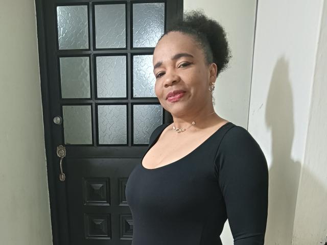 FernandoAndIsabella has medium brown skin, curly black hair in a bun, wearing a black long-sleeve top, pink lipstick, and a necklace, standing in front of a black door.