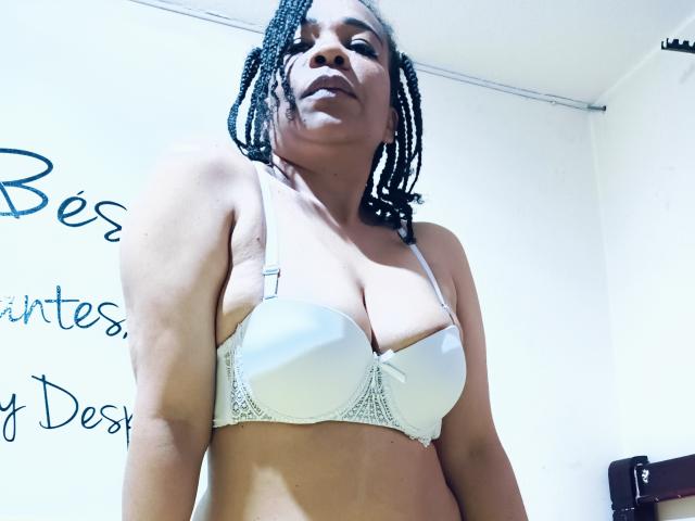 FernandoAndIsabella, dark-skinned woman with braided hair, wearing a white bra, showing her large tits, standing with a confident, slightly downward gaze.