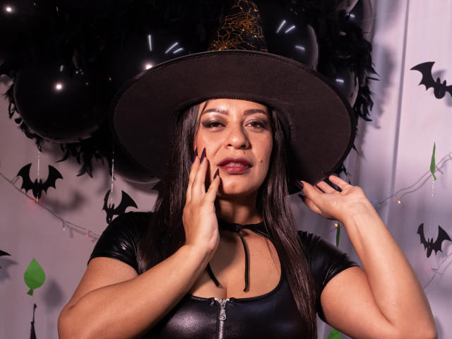 MiaCallie with dark brown hair, wearing a black witch hat, shiny black top, and red lipstick, touching her face, showing off her tits and ass in a Halloween-themed photo.