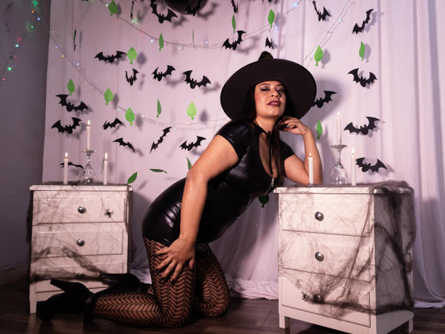 MiaCallie kneeling in black latex dress, hat, and fishnet stockings, showing her ass, with candles on marble nightstands, bat decorations, and a sexy, confident expression.