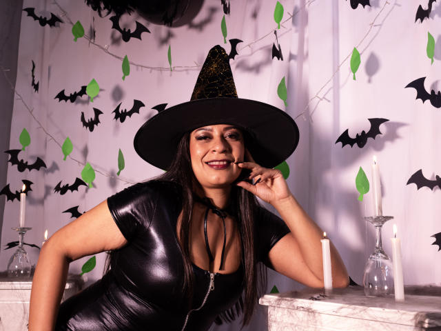 MiaCallie with long black hair, wearing a shiny black latex dress, black witch hat, and smiling, has her finger near her mouth, with candles and bat decorations.