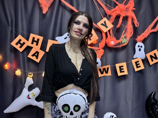 MilaLoona met lang bruin haar, een zwart shirt met losse bovenkant die haar borsten laat zien, met een schedelmasker in haar hand, een zwarte choker om, staand voor Halloween-decoratie..