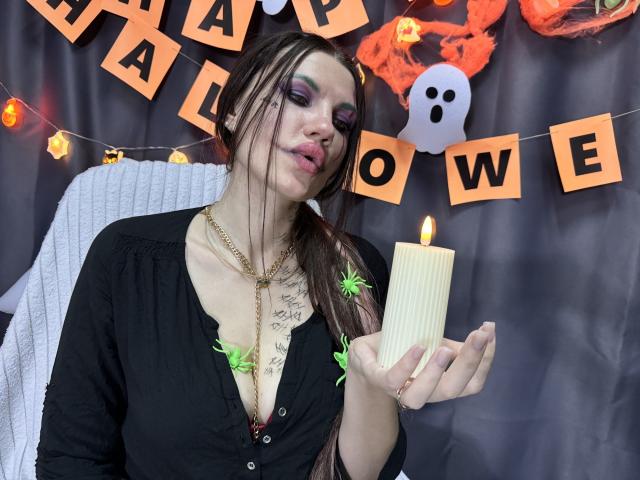 MilaLoona com cabelo trançado, maquiagem roxa nos olhos, blusa preta, colar, segurando uma vela, decorações de aranhas verdes, seios visíveis, peito tatuado, olhando para a câmera, tema de Halloween..