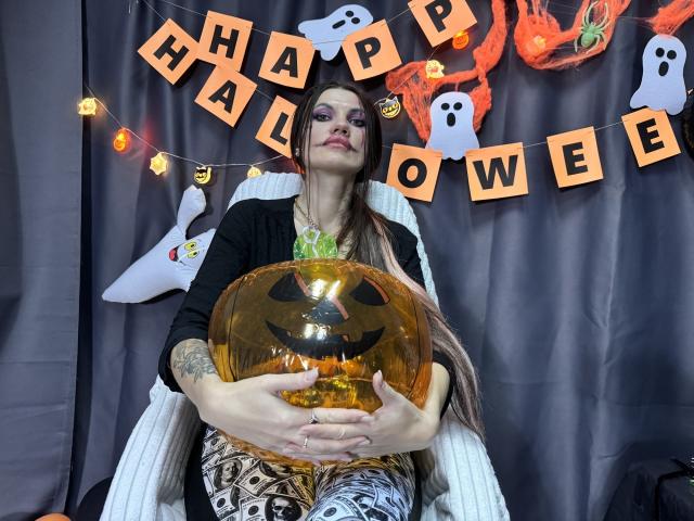 MilaLoona con cabello largo y castaño, maquillaje llamativo, un cárdigan negro, una blusa verde, pantalones estampados, sosteniendo una calabaza naranja con ojos negros, sentada en una silla blanca, con decoración de Halloween en el fondo..