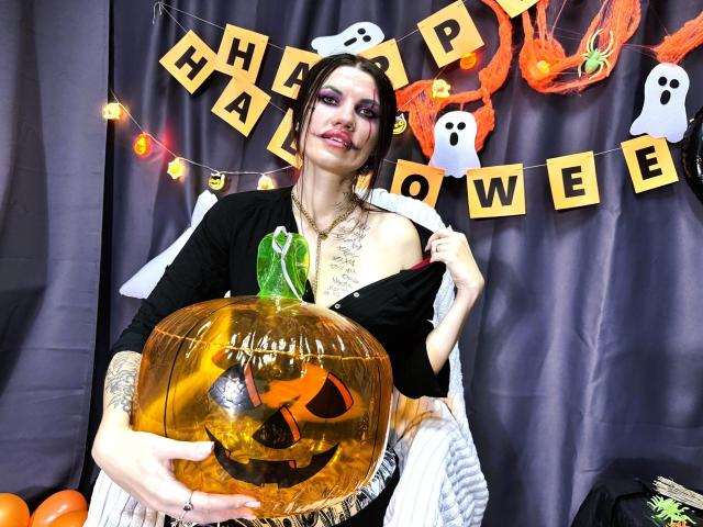 MilaLoona with purple eyeshadow, black top, and white dress, holding a glowing orange pumpkin with a carved jack-o'-lantern face, and a green stem. She has tattoos on her arm.